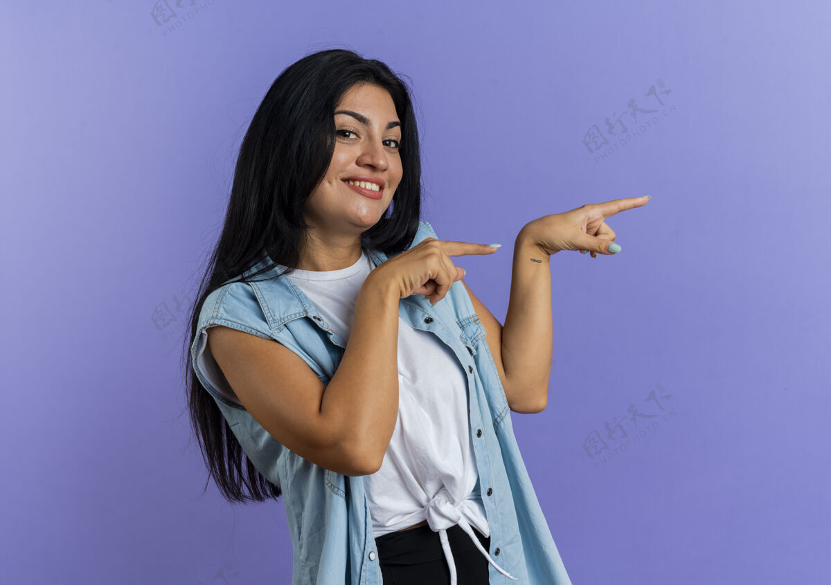 手微笑着的年轻白种女人用两只手指着侧面 看着隔离在紫色背景和复印空间上的相机背景点女人