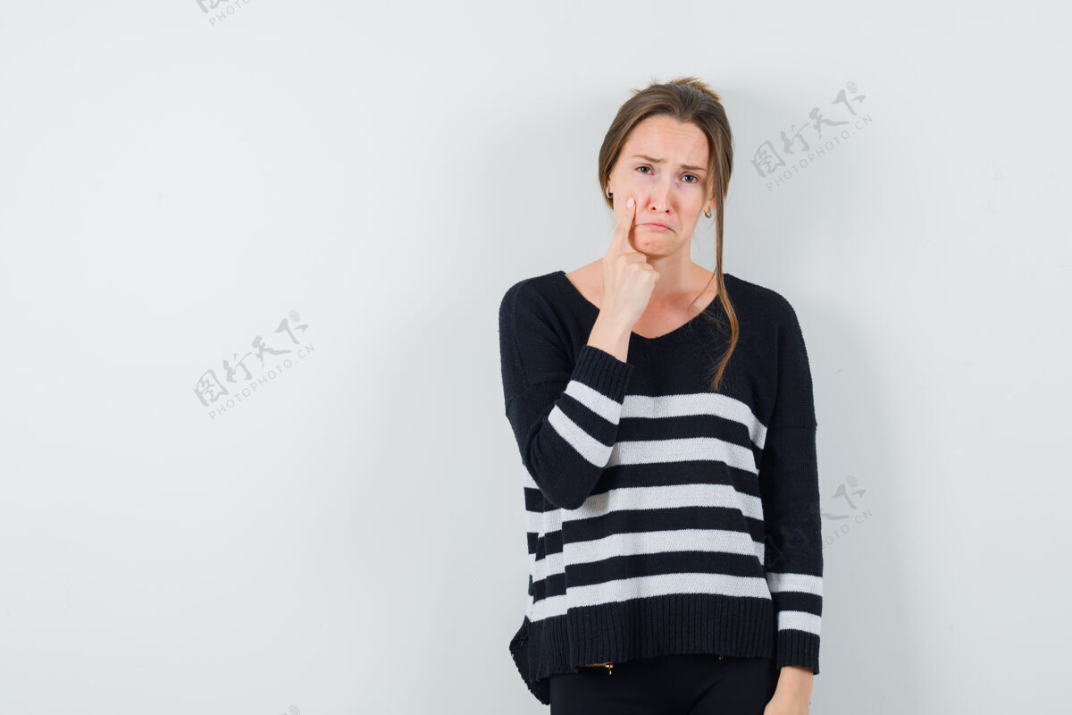 深色穿着条纹针织衫和黑色裤子的年轻女子用食指指着脸颊 看上去很沮丧女人自然目录