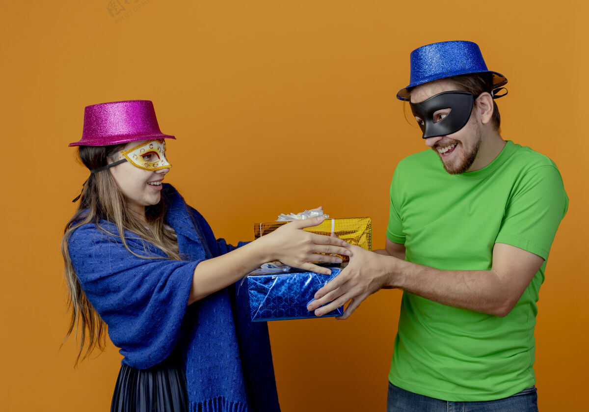蓝色高兴的年轻夫妇戴着粉红色和蓝色的帽子戴上化装眼罩看着对方拿着礼品盒隔离在橙色的墙上橙色年轻人情侣