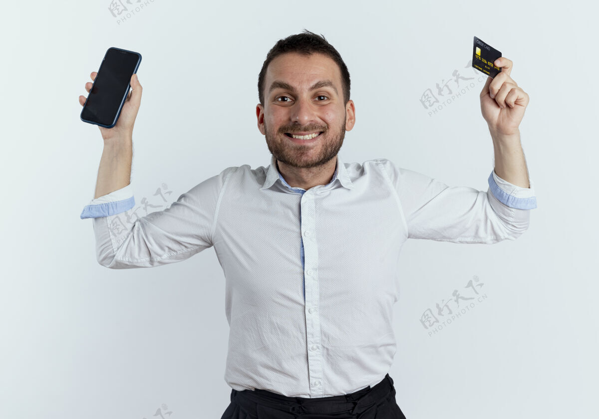 信用笑容可掬的帅哥举手拿着电话和信用卡隔离在白墙上持有年轻商人