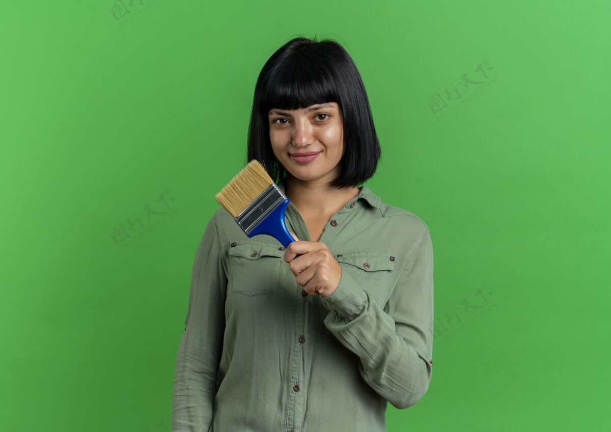 女士高兴的年轻黑发白种女人拿着油漆刷隔离在绿色背景与复制空间画笔黑发请拿着