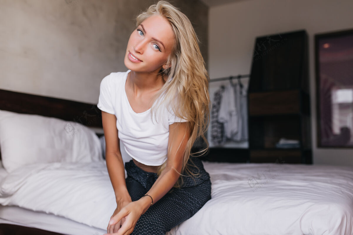 金发早上害羞的蓝眼睛女人在家里摆姿势穿着白色t恤的漂亮长发年轻女人坐在床上公寓人室内