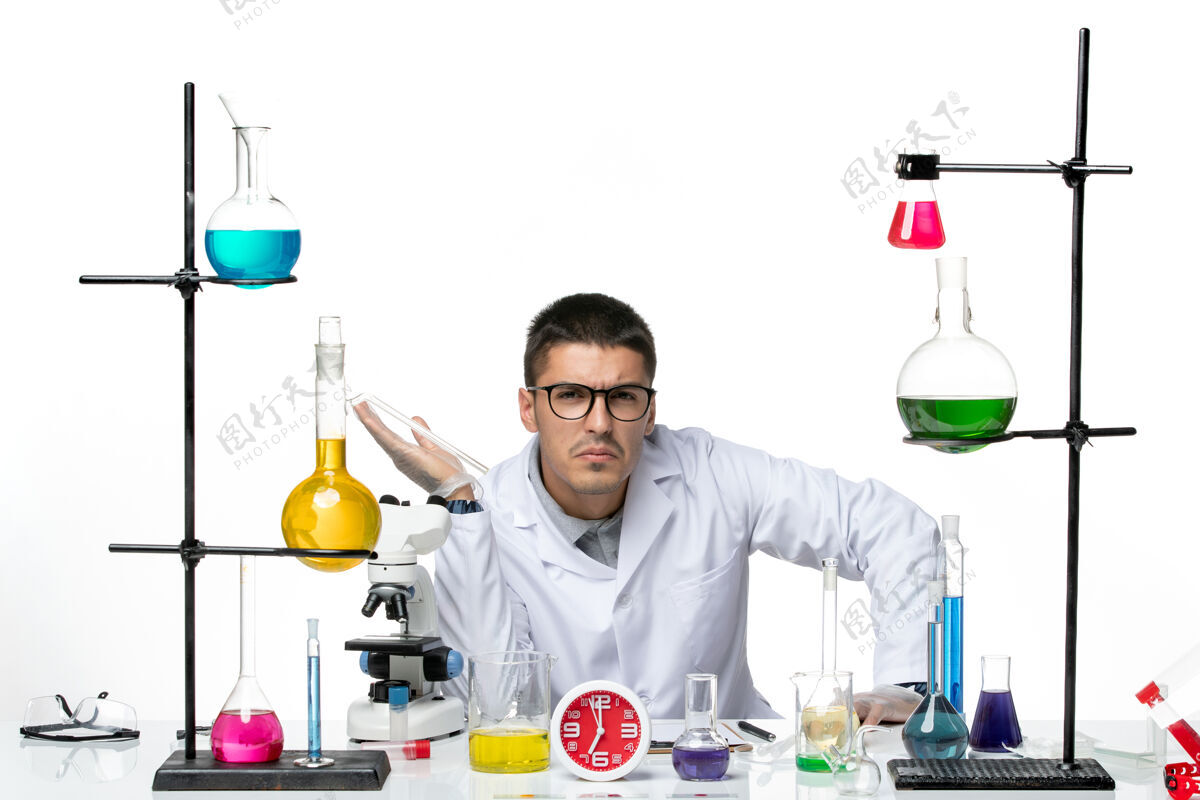 医学前视图穿着白色医疗服的男性化学家坐在白色背景病毒疾病科学实验室covid上 拿着不同的溶液实验室医生套装