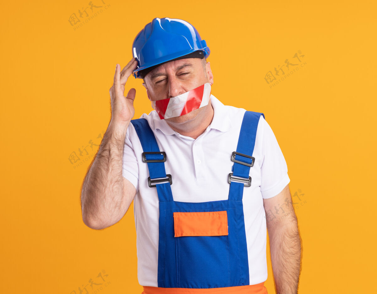 手一个穿着制服的白人成年建筑工人用胶带封住嘴巴 把手放在橘子树的头上封面人不愉快