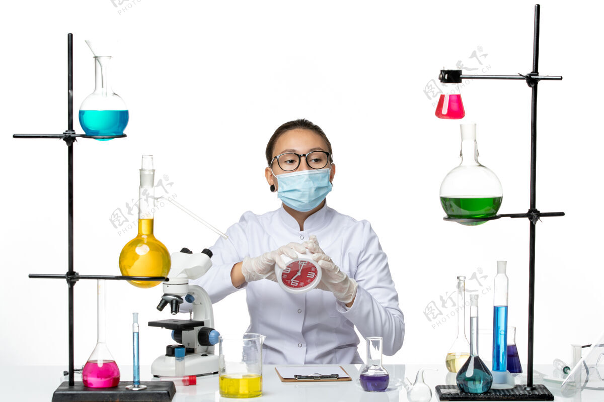 化学前视图：身穿医疗服的女化学家戴着面具 白色办公桌上拿着时钟病毒实验室化学科维德飞溅飞溅持有瓶子