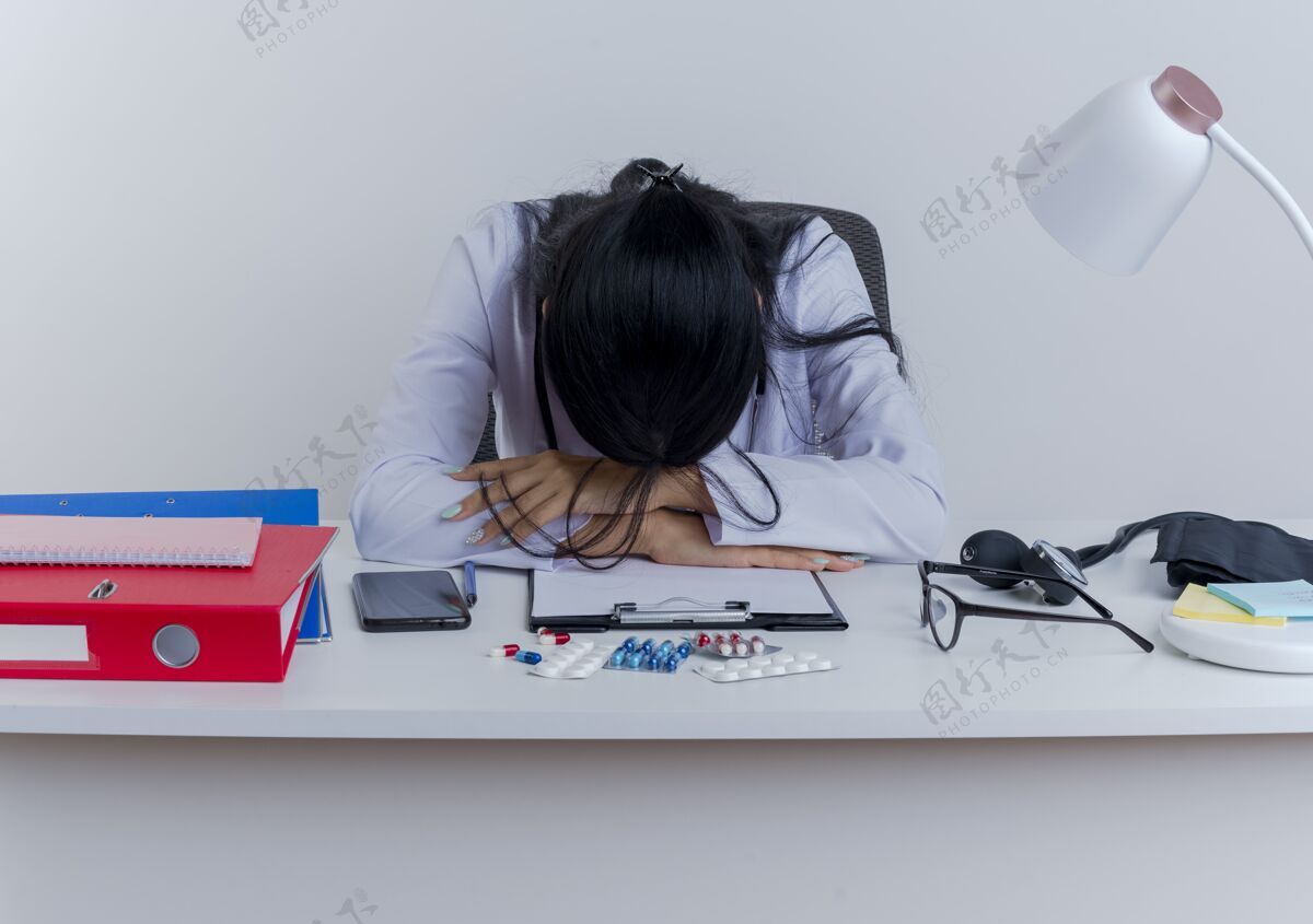 女人疲惫的年轻女医生穿着医用长袍和听诊器坐在办公桌旁 手放在办公桌上 头放在手上坐着成人工具
