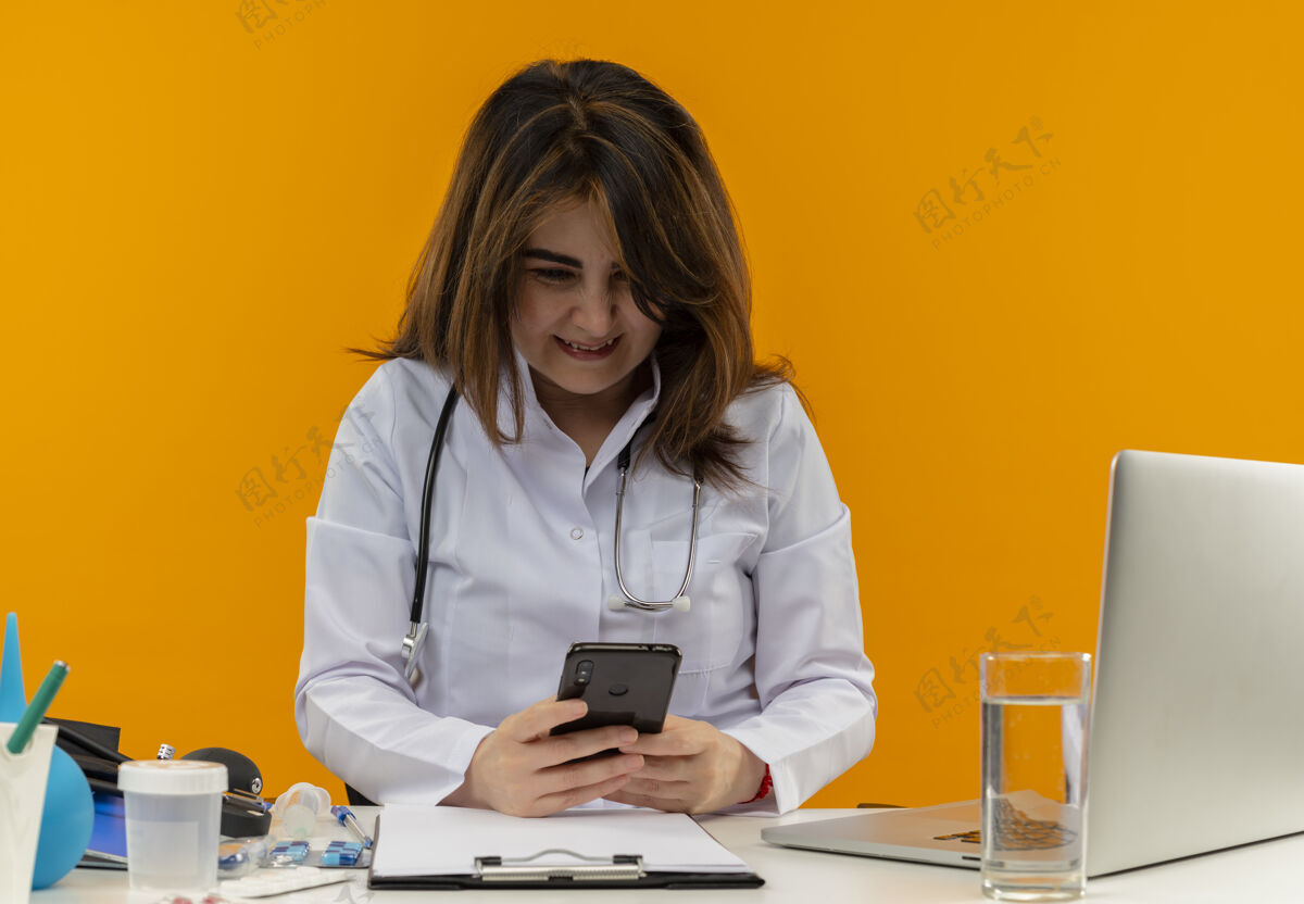 手机微笑的中年女医生穿着医用长袍和听诊器坐在办公桌旁 用医疗工具剪贴板和笔记本电脑用手机隔离穿着女性医生