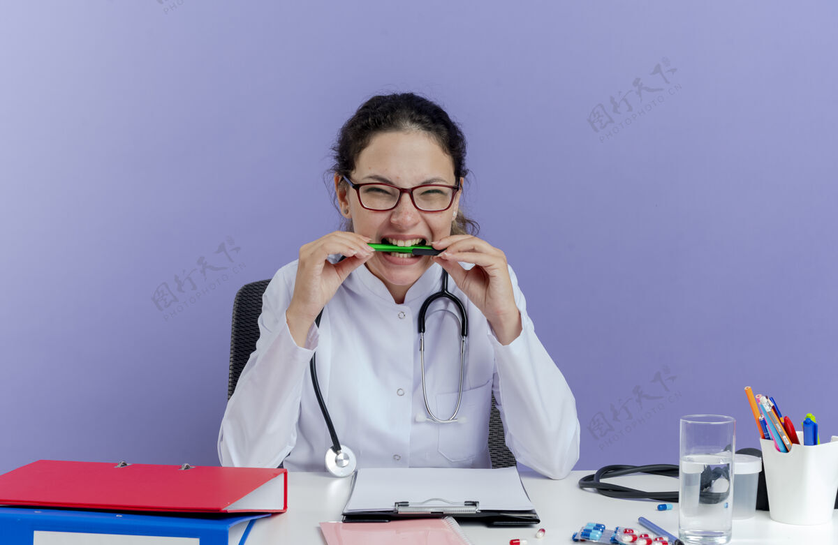 年轻人压力重重的年轻女医生穿着医用长袍和听诊器坐在办公桌旁 手里拿着医疗工具 看起来像是一只被隔离的咬着的钢笔女性医生听诊器
