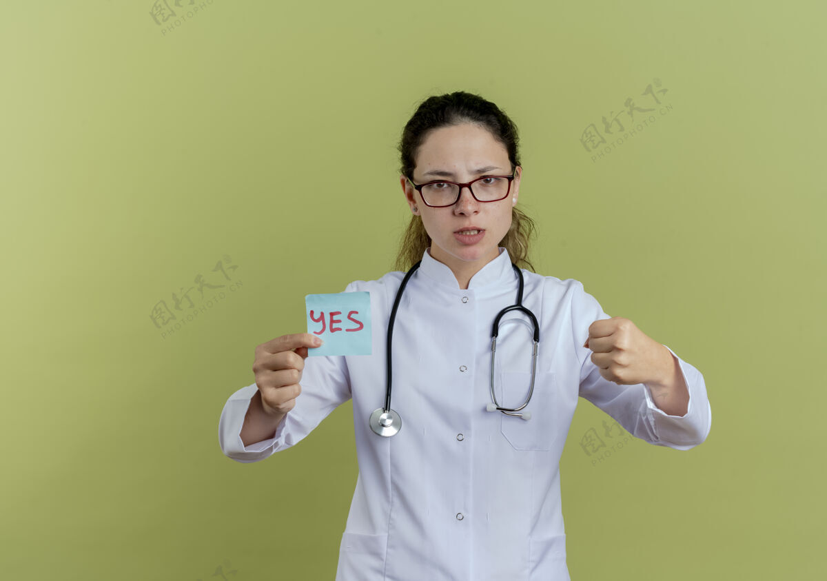 年轻严格的年轻女医生 穿着医用长袍 戴着听诊器 戴着眼镜 拿着纸条 显示着拳头眼镜拳头人
