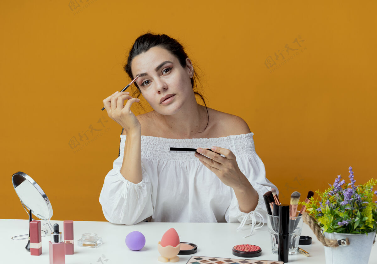 刷子自信美丽的女孩坐在桌子上 用化妆工具拿着化妆刷和眼影调色板 看起来在橙色的墙上女人工具坐