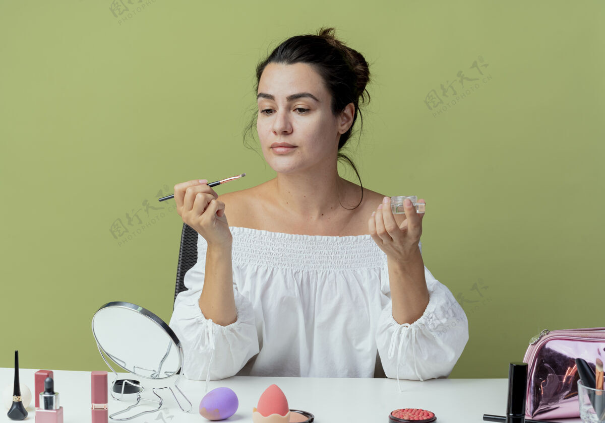 桌子美丽的女孩坐在桌子上用化妆工具准备应用眼影和化妆刷隔离在绿色的墙上刷子涂抹坐