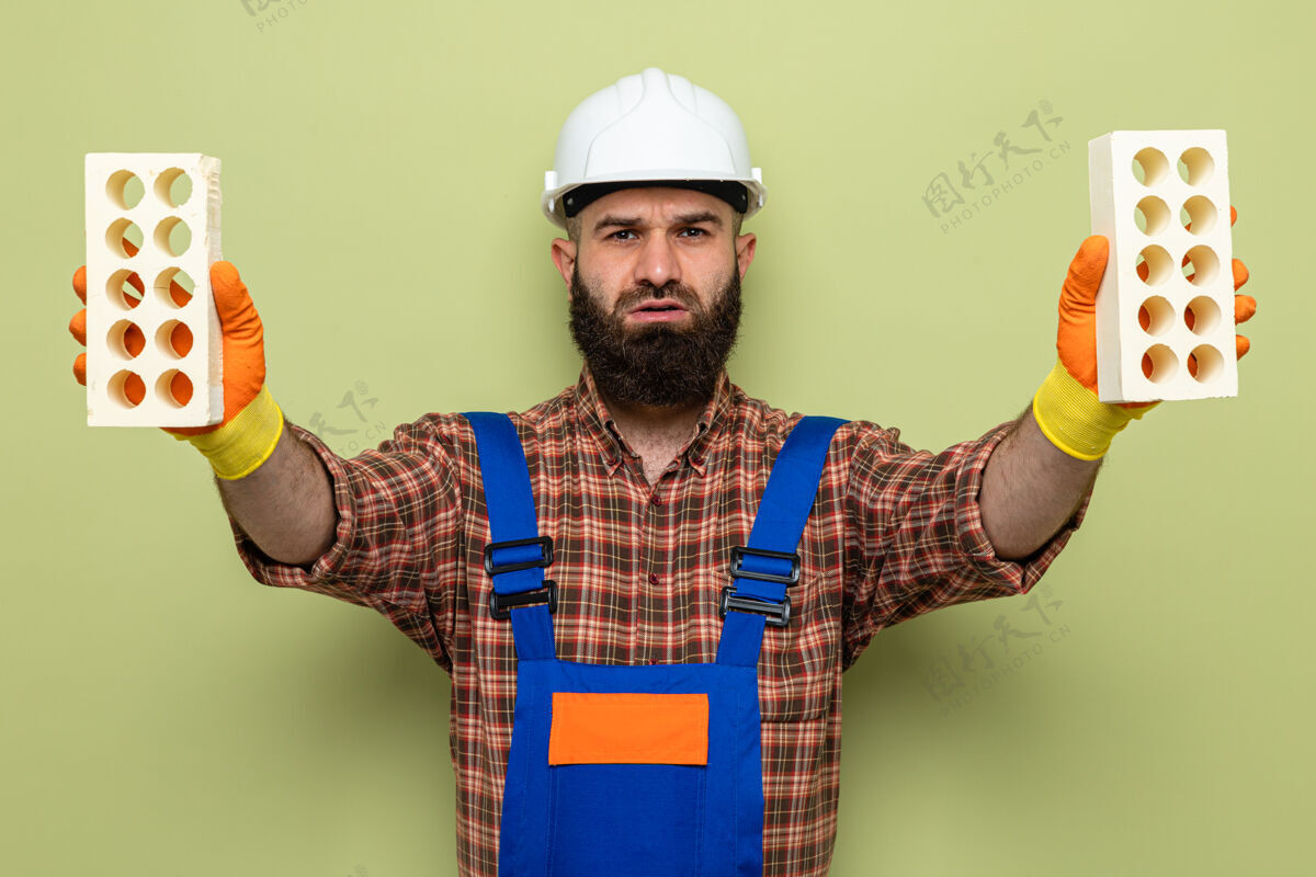 砖块留着胡子的建筑工人穿着建筑制服 戴着安全帽 戴着橡胶手套 拿着砖块 严肃地看着摄像机站在绿色的背景上胡须建筑工人手套