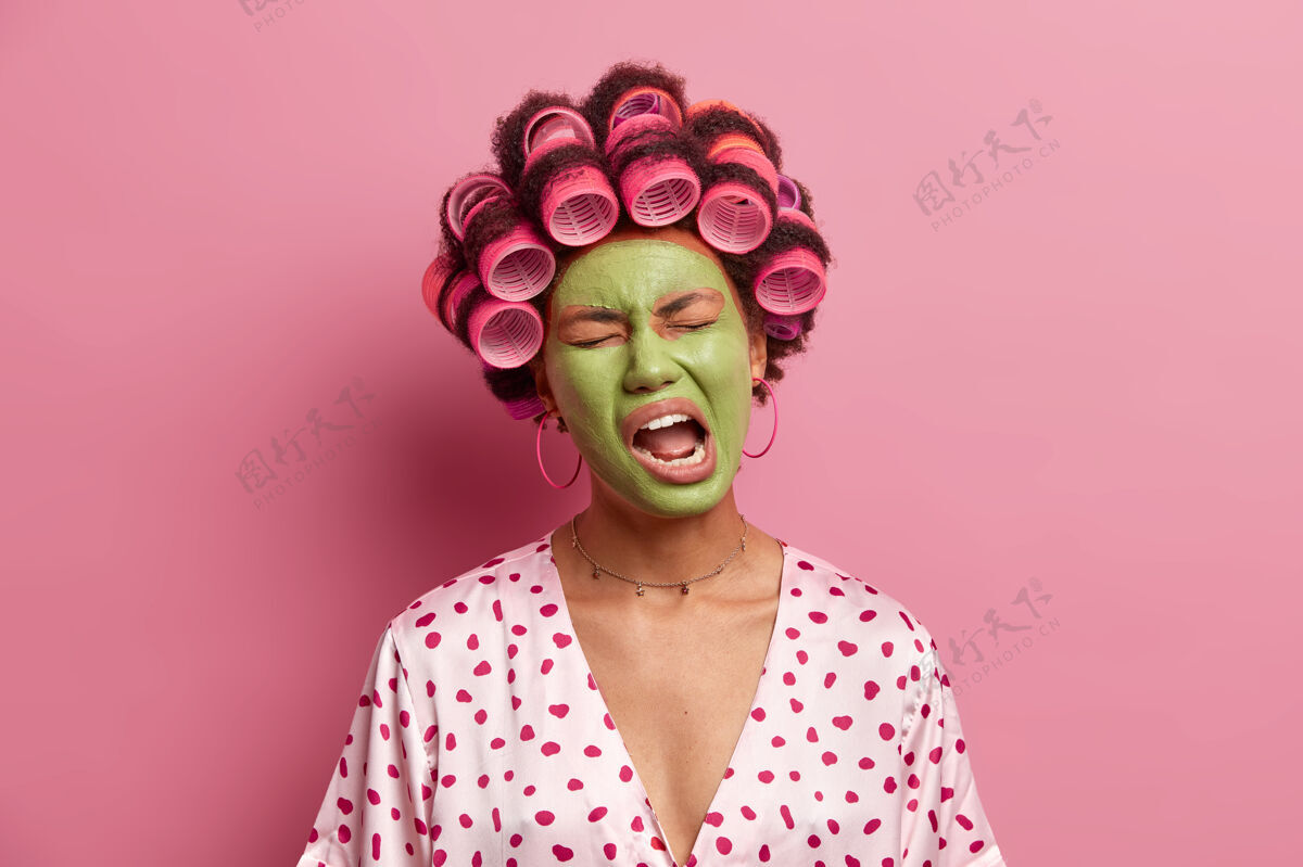 粘土不高兴的少数民族妇女张着嘴 感到疲劳 打哈欠 戴着绿色的口罩 穿着休闲的家装 隔离在粉红色的脸上面部护理和健康理念不高兴长袍宠爱