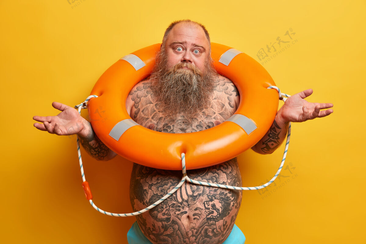 进退两难不确定的大胡子男人 肚子上有纹身 双手侧着 感到怀疑和犹豫 站在橙色的救生圈里 学习如何游泳 孤立在黄色的墙上游泳的时间到了男人人白种人