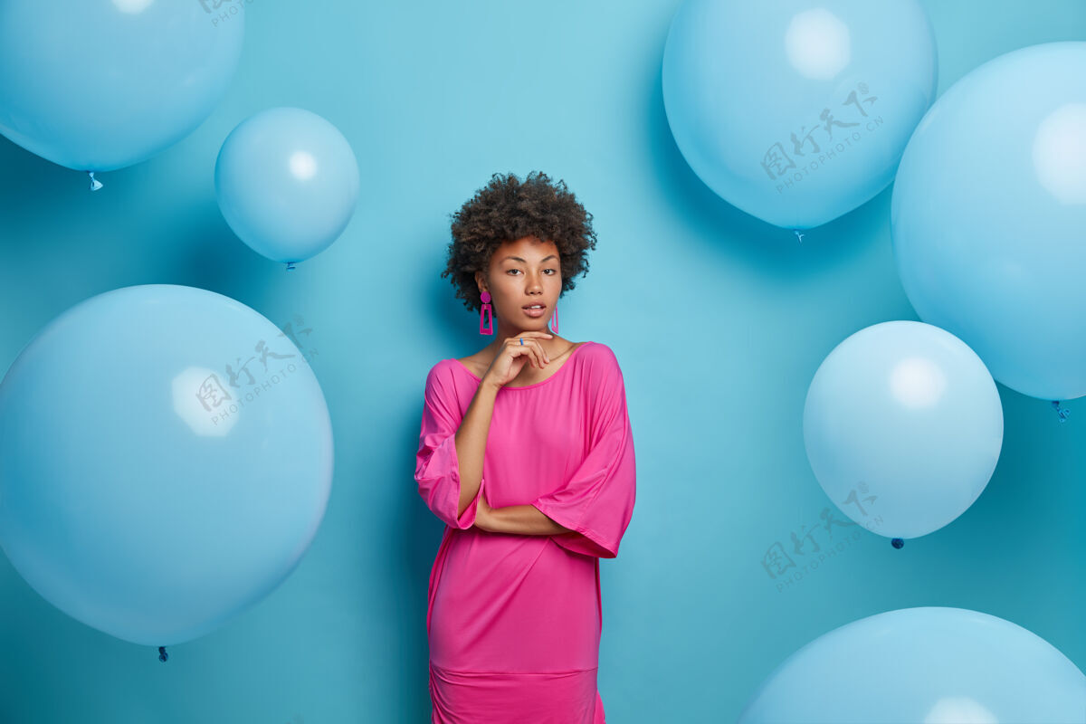 穿着严肃自信的女士的肖像托着下巴直视 穿着玫瑰色的裙子 对着蓝色墙壁上孤立的派对上的气球摆姿势穿着优雅衣服的迷人的女模特下巴节日氦气