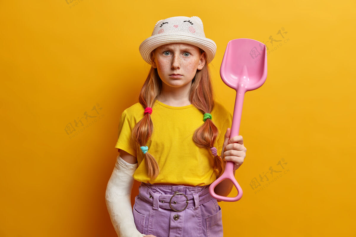青少年严厉严肃的红发女孩横拍在沙滩上玩 拿着粉色塑料铲 穿着时髦的衣服 做了危险运动后在石膏上绷带断了胳膊 隔离在黄色的墙上绷带铲子伤害
