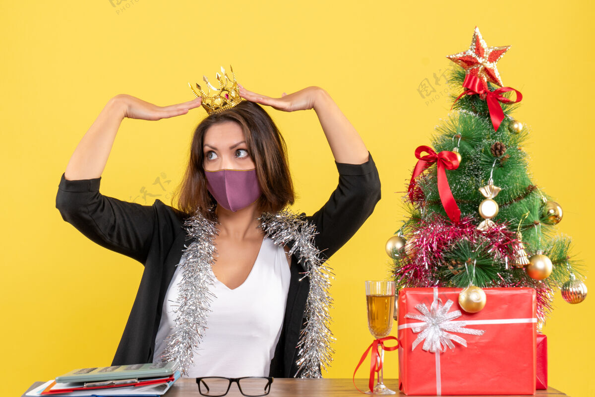 医疗圣诞节的心情与美丽的女士穿着西装戴着皇冠与医疗面具和手持礼物在黄色的办公室头发美女成人