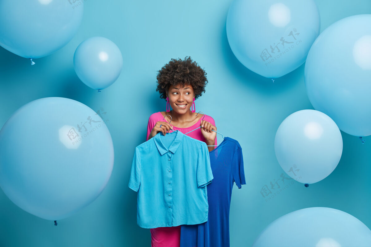 高兴时尚女人在两件衣服中挑选 衣架上拿着蓝色的连衣裙和衬衫 想穿什么好看就要在社团聚会上若有所思地站在室内一边室内购买衣服