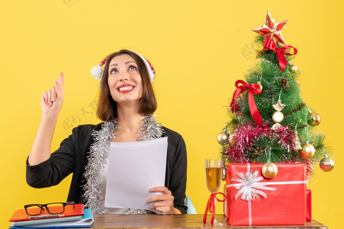 快乐一位身着西装 戴着圣诞老人帽 戴着新年装饰品 面带微笑的商务女士独自一人指着上方 坐在办公室里一张桌子旁 桌子上放着圣诞树帽子指着美丽