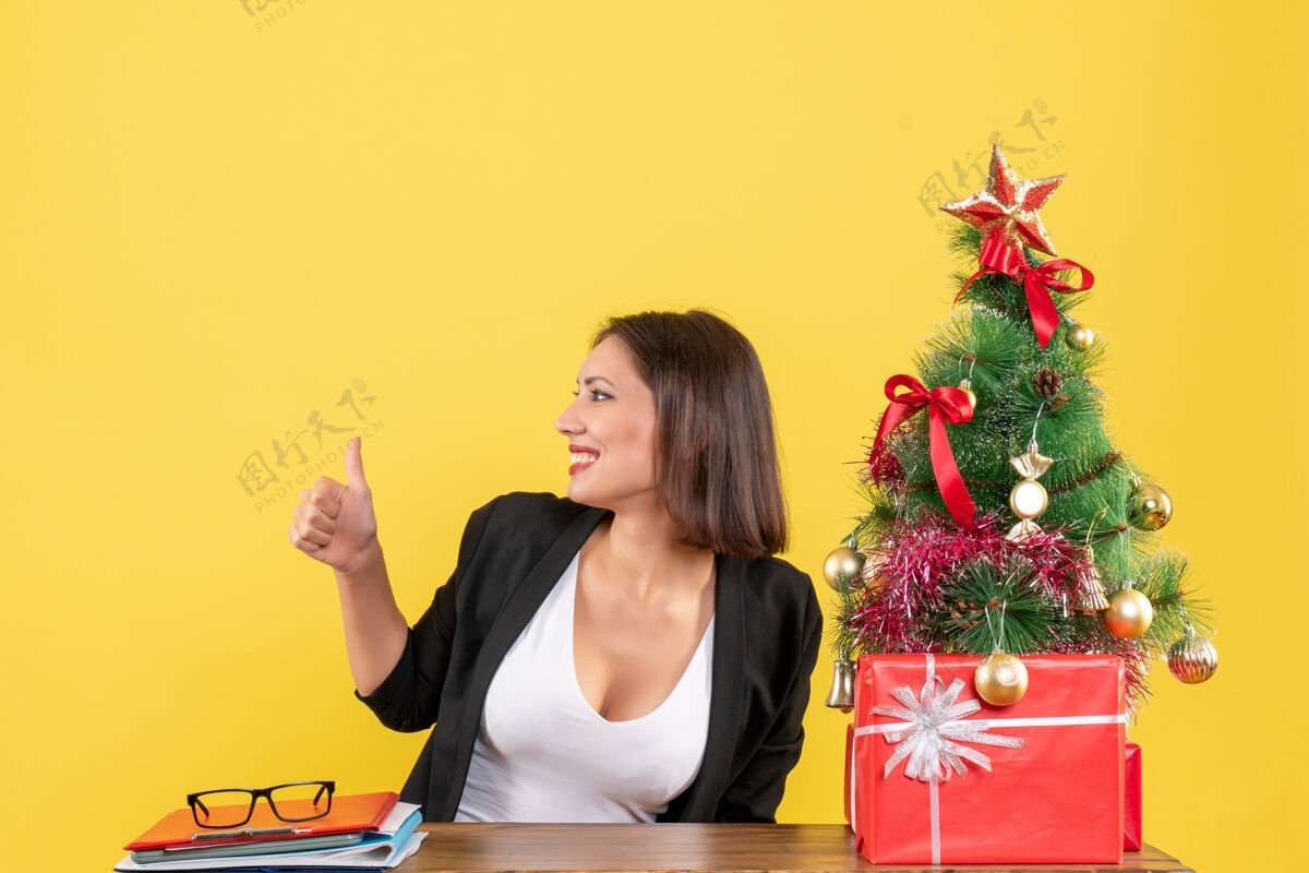 年轻女子在黄色的办公室里 一位穿着西装的年轻女士在装饰好的圣诞树旁做着“ok”的手势好圣诞手势