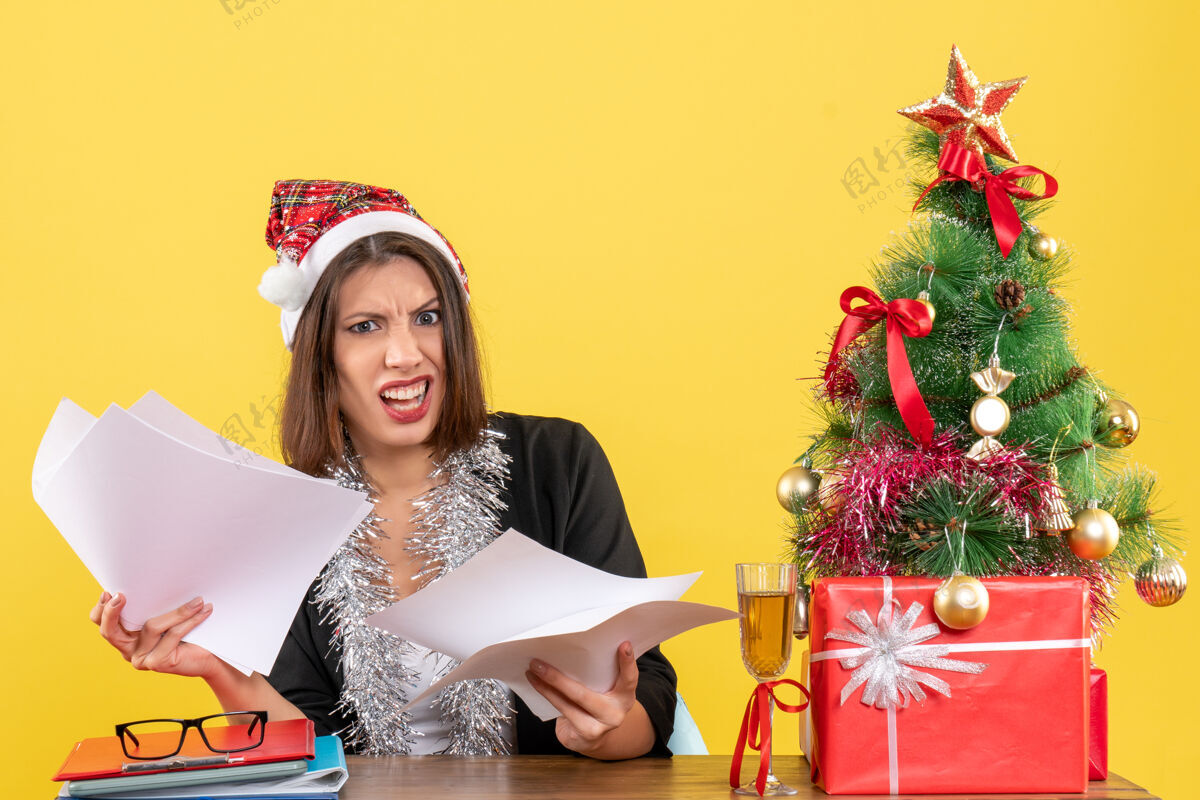文件情绪混乱的商务女士穿着西装 戴着圣诞老人的帽子和新年装饰品 手里拿着文件 坐在一张桌子旁 桌子上放着圣诞树圣诞老人套装树