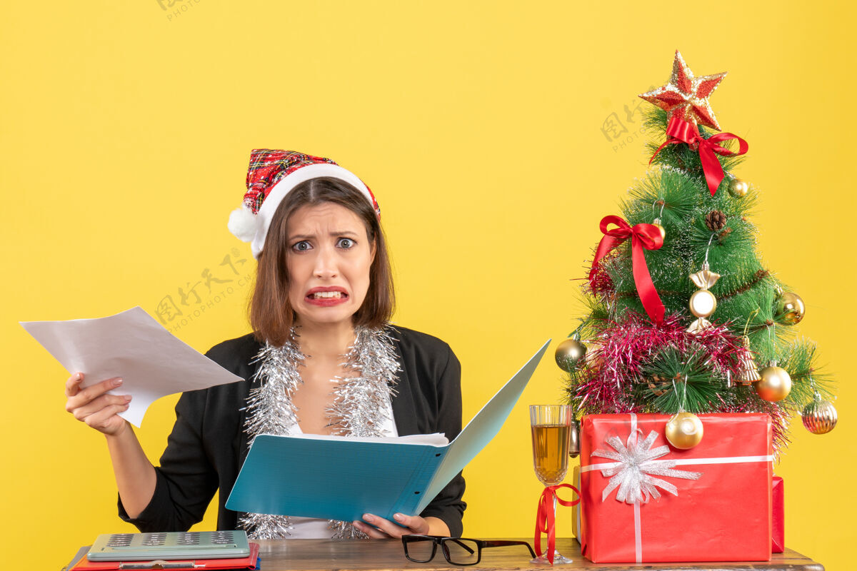 新郎迷茫迷人的女士穿着西装 戴着圣诞老人的帽子和新年的装饰品 手里拿着文件 在黄色的办公室里与世隔绝抱年圣诞老人