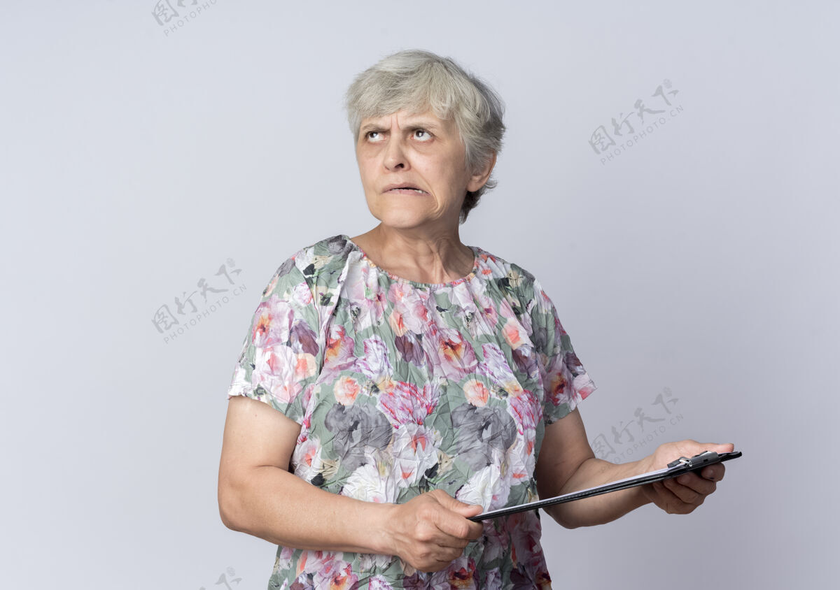 起来困惑的老妇人拿着剪贴板 孤零零地看着白色的墙上剪贴板困惑老人