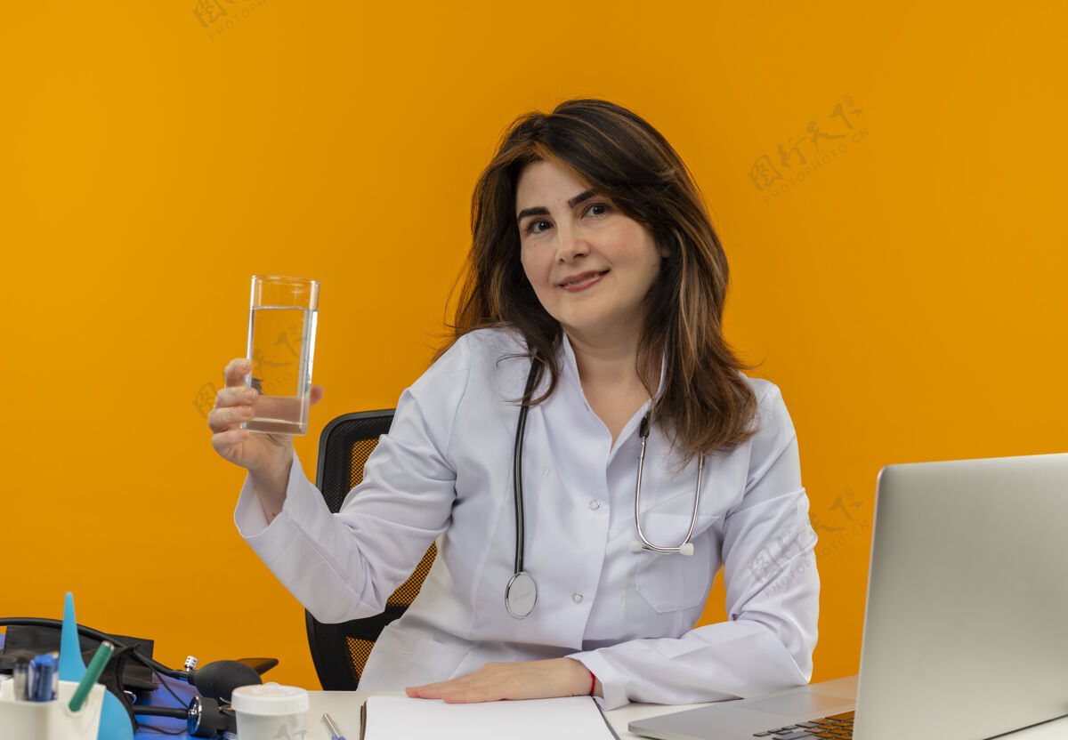水高兴的中年女医生穿着医用长袍和听诊器坐在办公桌旁 手里拿着医疗工具剪贴板和笔记本电脑 手放在办公桌上 手里拿着一杯水笔记本电脑人桌子