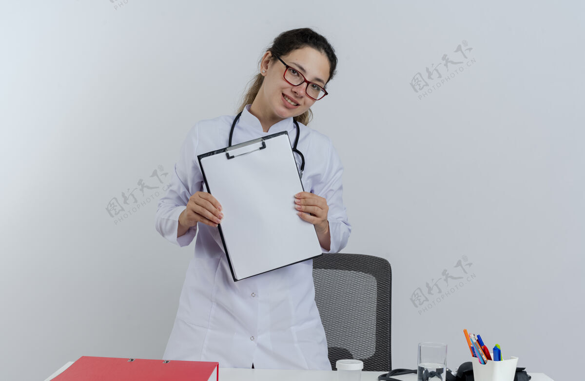医生面带微笑的年轻女医生穿着医用长袍 戴着听诊器 戴着眼镜 站在桌子后面 带着医疗工具 看上去与世隔绝表演剪贴板成人
