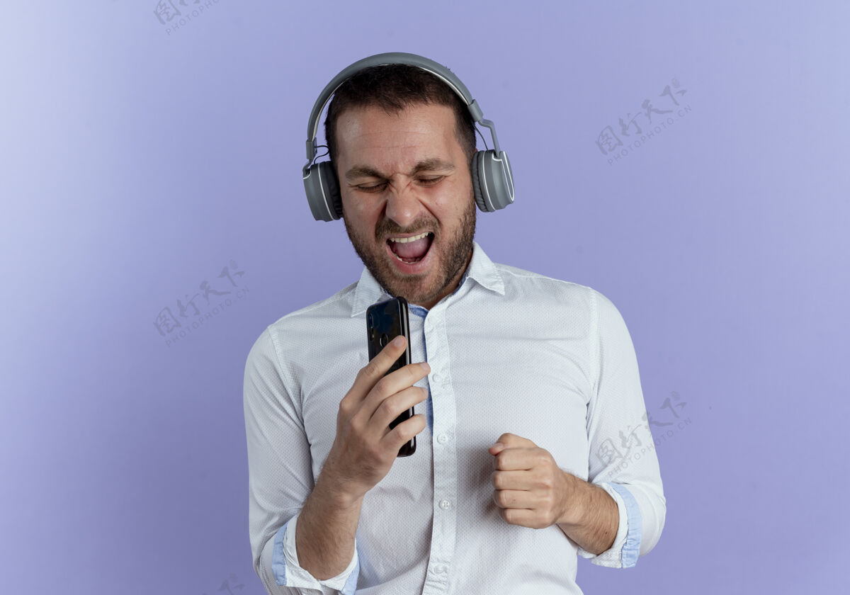 耳机戴着耳机的快乐帅哥拿着电话假装在紫色的墙上孤立地唱歌男人紫色快乐