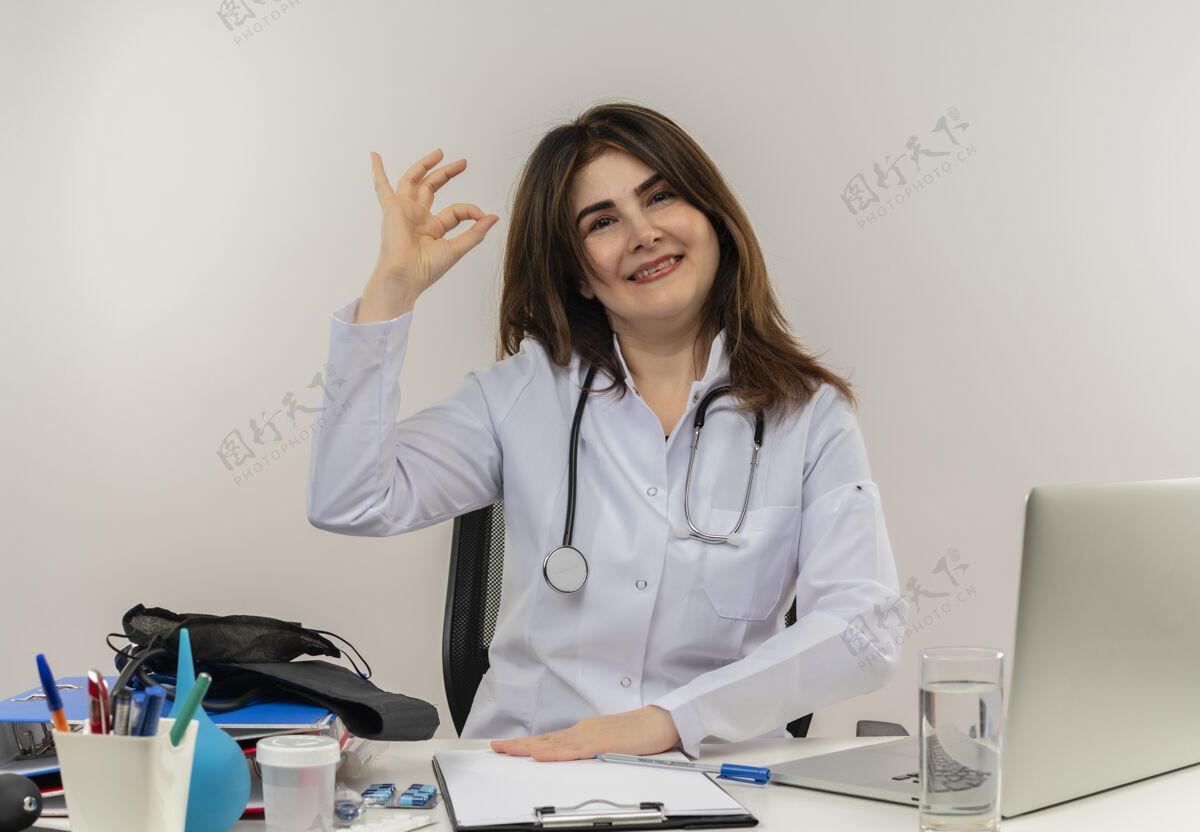 办公桌微笑的中年女医生穿着医用长袍和听诊器坐在办公桌前 拿着医疗工具剪贴板和笔记本电脑做着ok标志隔离穿着女性可以