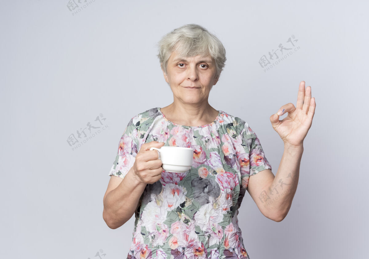 拿着高兴的老太太拿着杯子 手势ok的手势孤立在白墙上老人手势女人