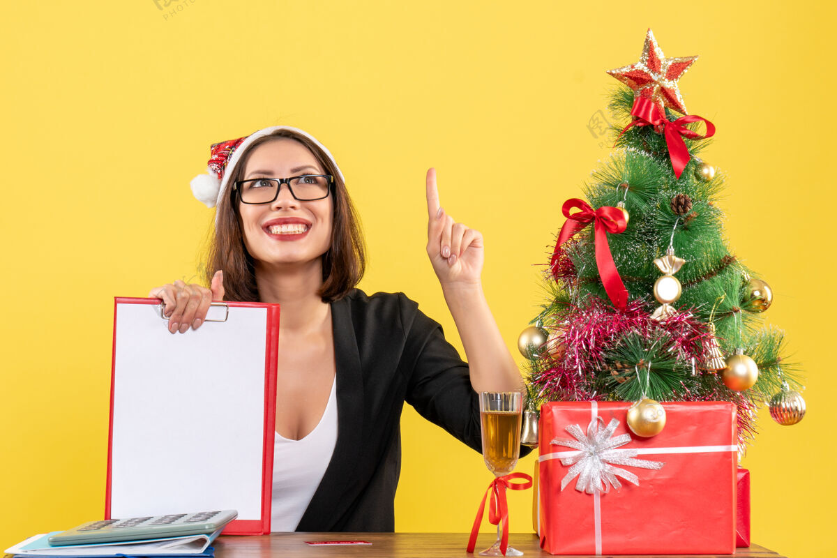 西装迷人的女士穿着西装 戴着圣诞老人的帽子 戴着一副眼镜 展示着文件指着办公室里的黄色隔离带微笑漂亮向上