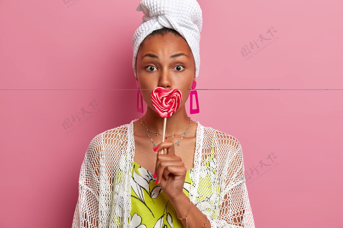 女人可爱健康的女人用明亮的心形棒棒糖捂住嘴 去满足她的甜食 穿着家装和珠宝 隔离在粉红色的墙上女人用美味的糖果棒棒糖风景室内
