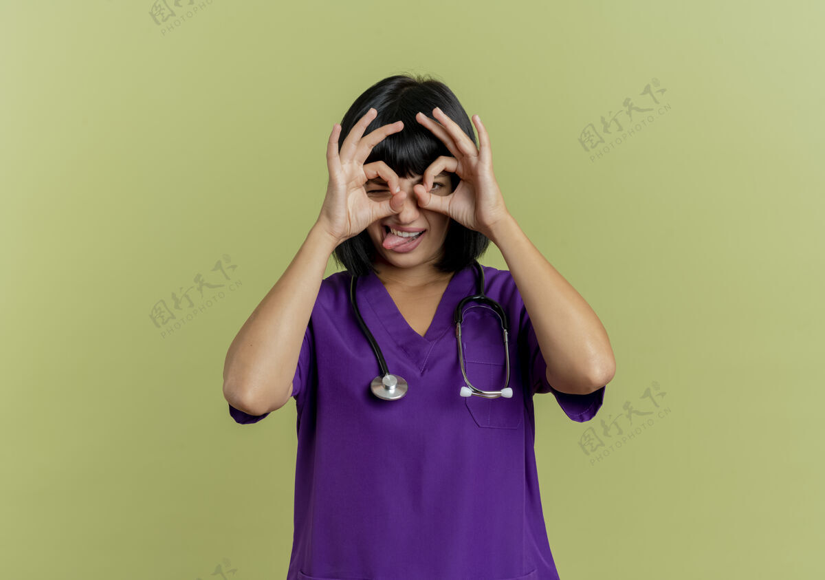 舌头快乐的年轻黑发女医生穿着制服 带着听诊器 从橄榄绿背景上的手指中探出舌头 并留有复印空间女性医生绿色