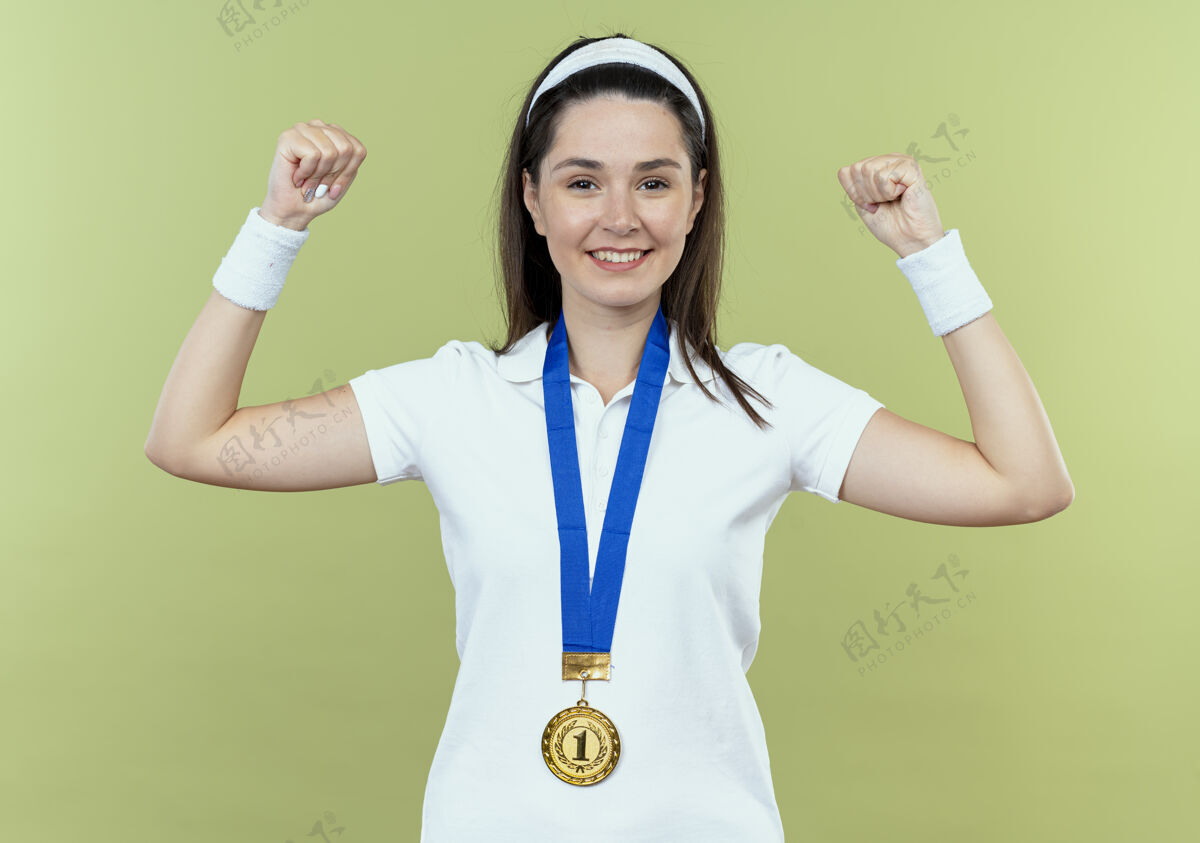 健身戴着头巾 脖子上戴着金牌的年轻健身女士举起拳头 站在灯光的背景下 脸上洋溢着幸福的笑容 看上去很自信奖牌站立微笑