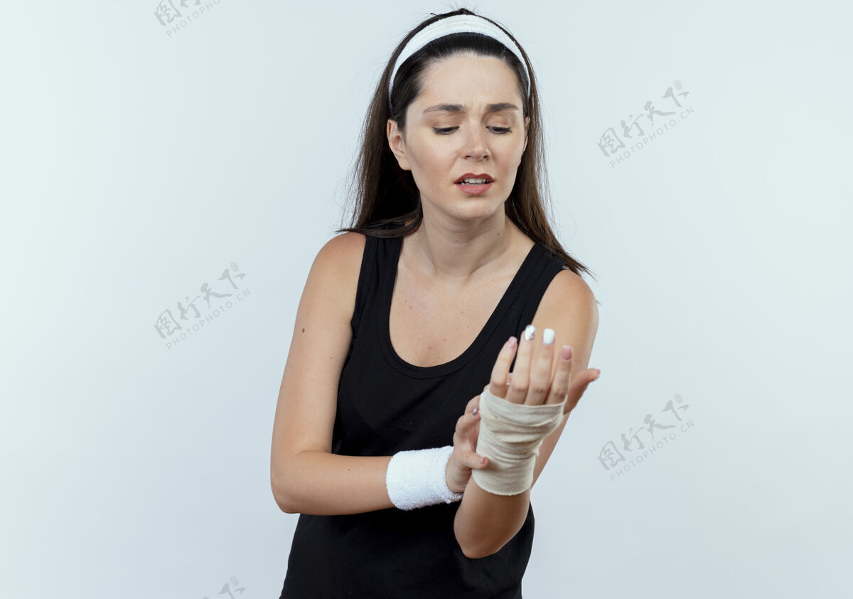 手腕戴着头巾的年轻健身女士站在白色背景下看着她缠着绷带的手腕感到疼痛绷带年轻头带