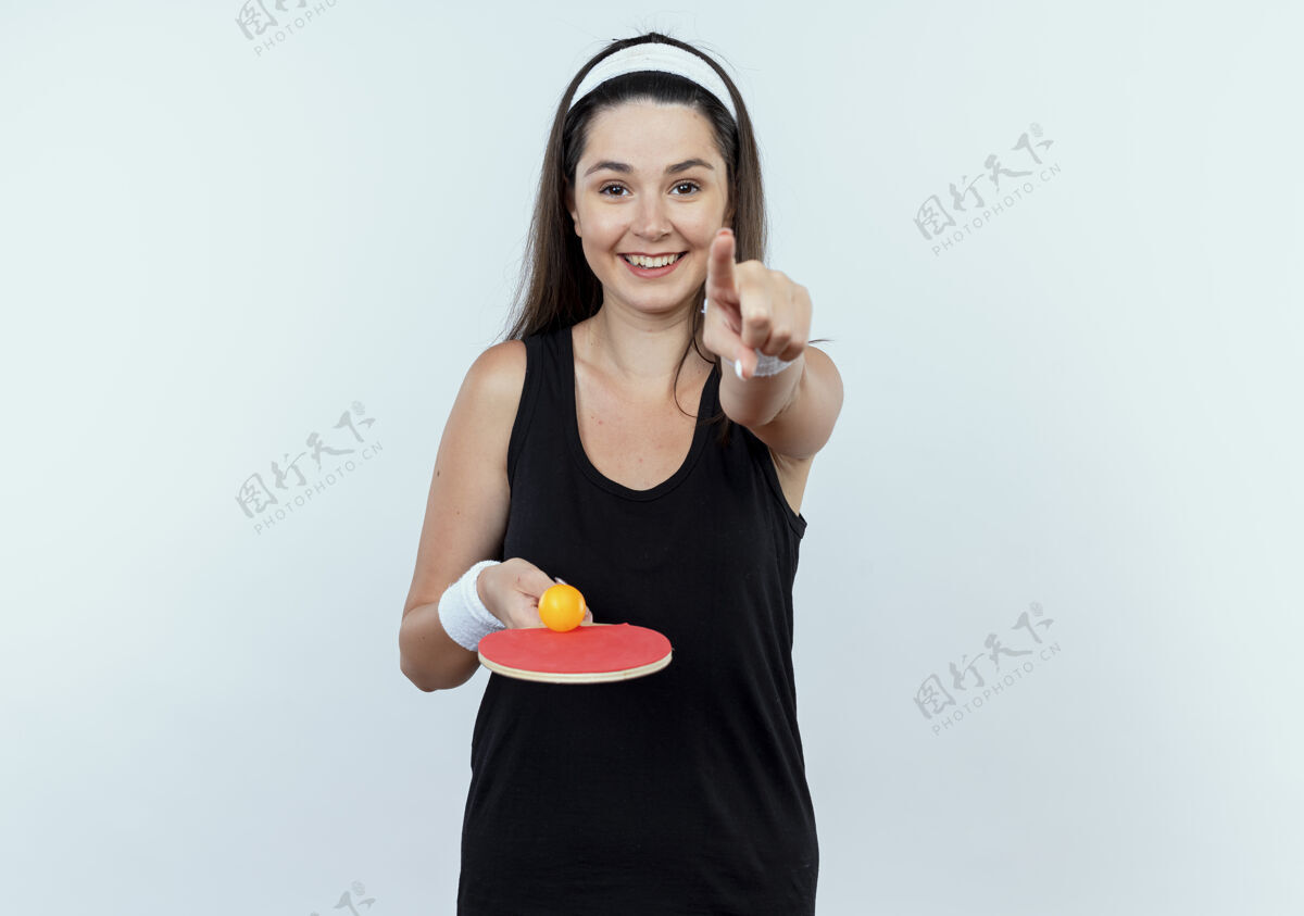 桌子戴着头巾 拿着球拍和乒乓球的年轻健身女子 手指对着镜头 站在白色背景上 欢快地微笑着网球年轻人欢呼