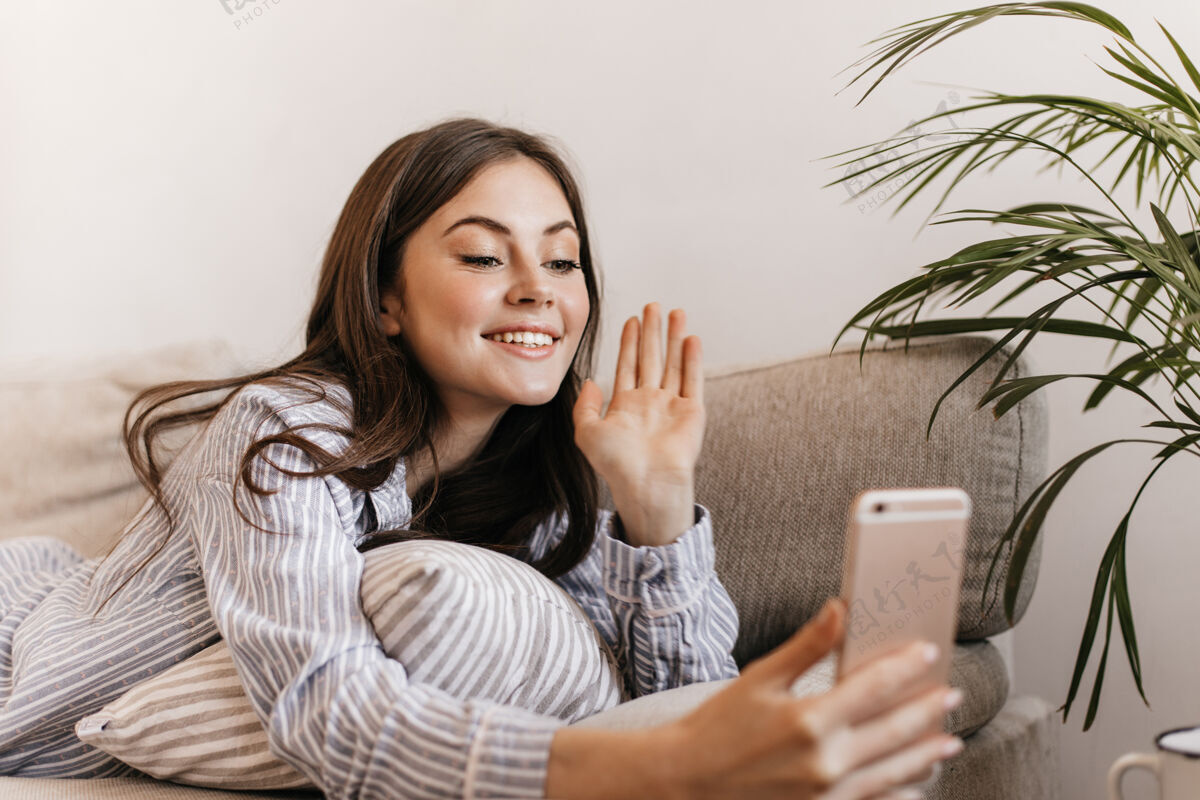 沙发一个穿着条纹衬衫的快乐女人躺在沙发上 通过电话里的视频聊天成人粉色房间