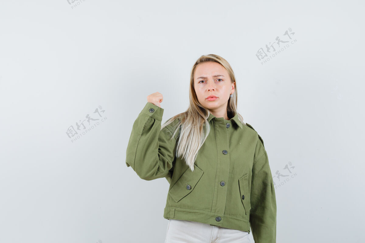 夹克身着绿色夹克 牛仔裤 表情严肃的年轻女性展示着她的手臂力量 正面照年轻微笑水疗