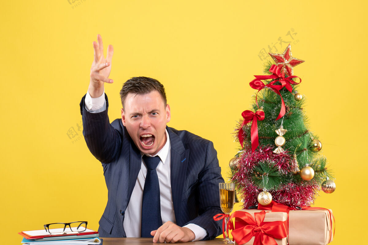 喊叫前视图愤怒的男子一边大喊大叫 一边坐在圣诞树和黄色背景上的礼物旁边的桌子上行政人员办公室愤怒的男人