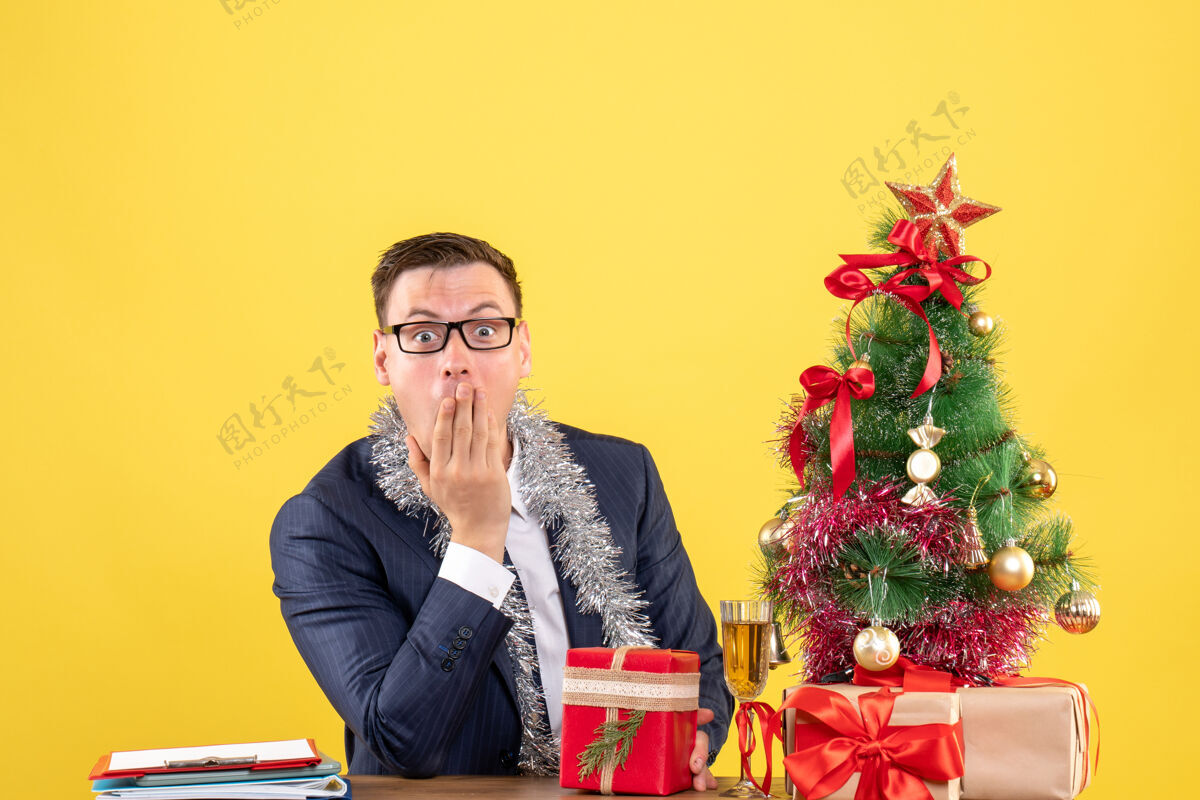 成人前视图惊奇的男人把手放在嘴边 坐在圣诞树旁的桌子上 呈现黄色背景背景人导演