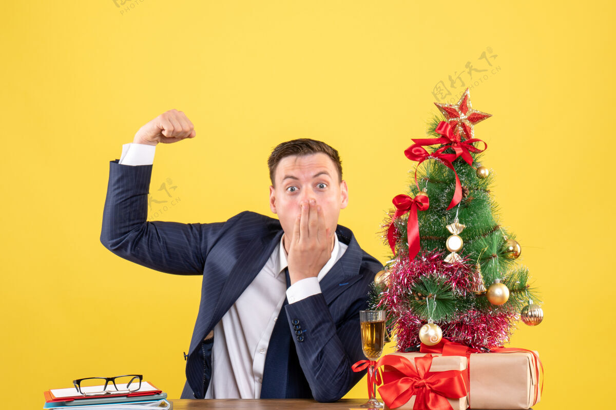 坐着前视图令人惊讶的男子显示肌肉坐在圣诞树附近的桌子上 并提出黄色背景肌肉办公室前面