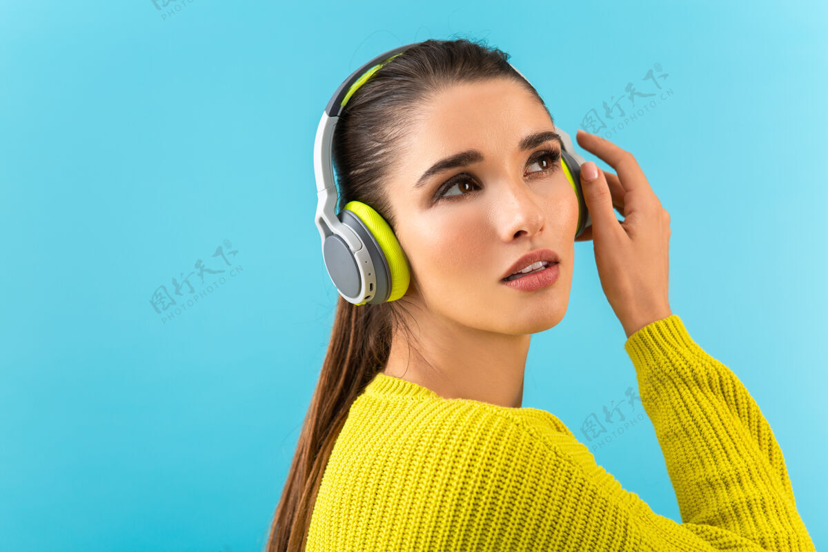 服装迷人时尚的年轻女子戴着无线耳机听音乐快乐地穿着黄色针织毛衣五颜六色的时尚造型隔离在蓝色的墙上休闲欢乐耳机
