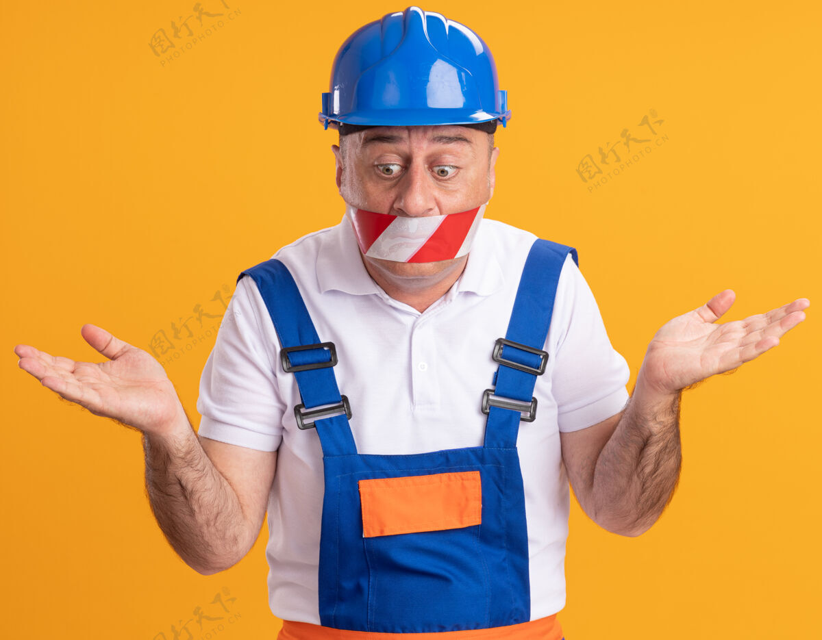 衣服焦虑的成年建筑工人穿着制服 用胶带捂住嘴巴 双手张开 看着橙色的墙壁建筑工持有脸