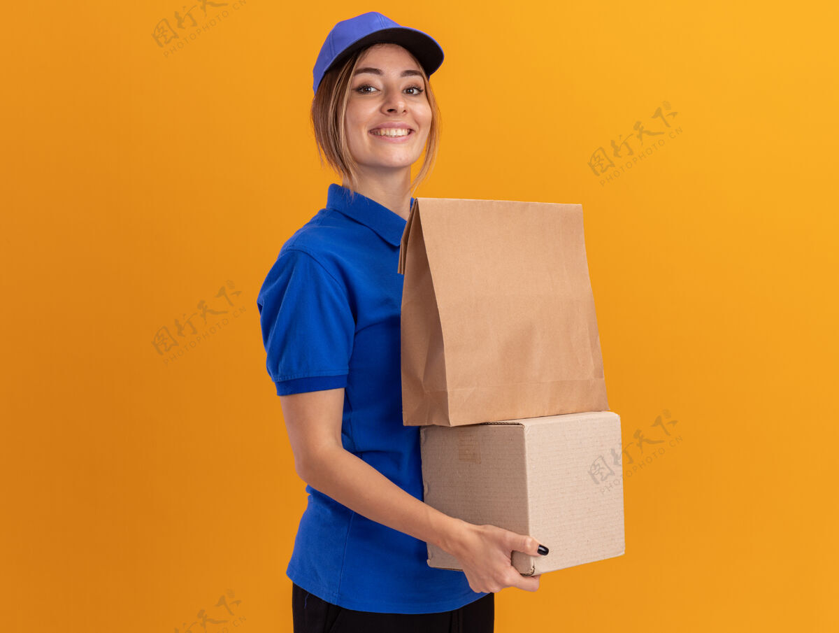 人身着制服 面带微笑的年轻漂亮女送货员把纸包放在隔离在橙色墙上的名片盒上制服举行感觉