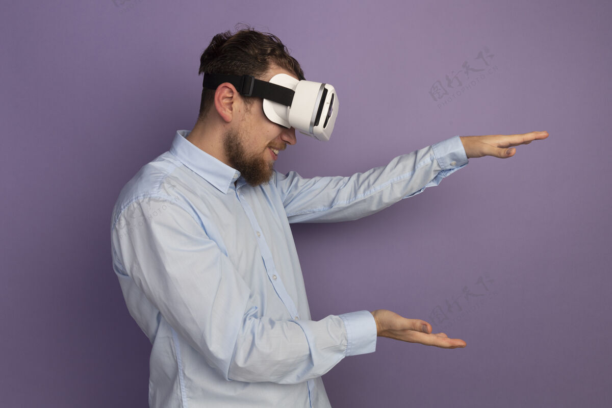男人面带微笑的金发帅哥戴着虚拟现实耳机假装拿着紫色墙上孤立的东西东西帅哥耳机