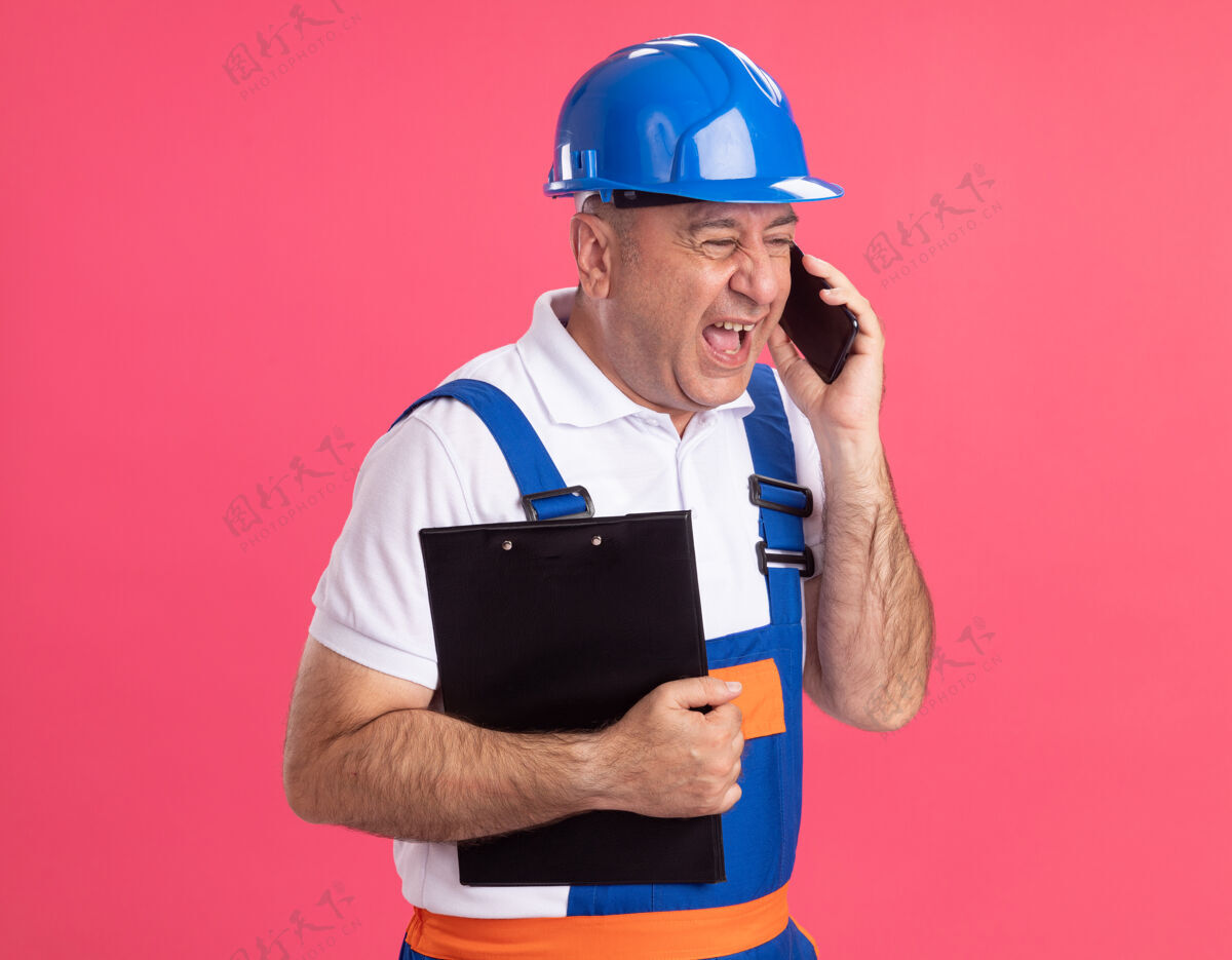 剪贴板恼怒的穿制服的成年建筑工人拿着剪贴板 隔着粉红色的墙上讲着电话脸公民电话