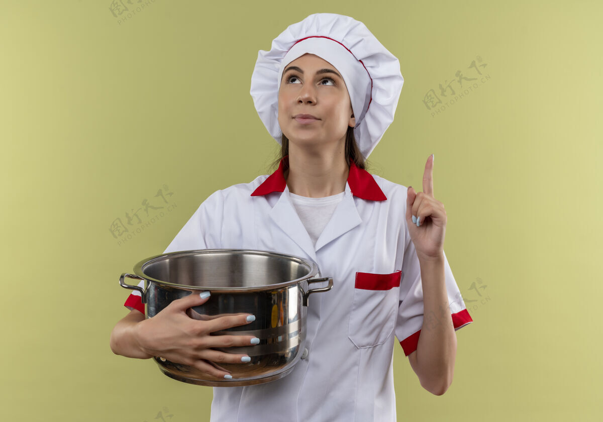 请身着厨师制服的年轻快乐的白人厨师女孩拿着锅子 在绿色背景上孤立地指了指 还有复印空间厨师年轻点