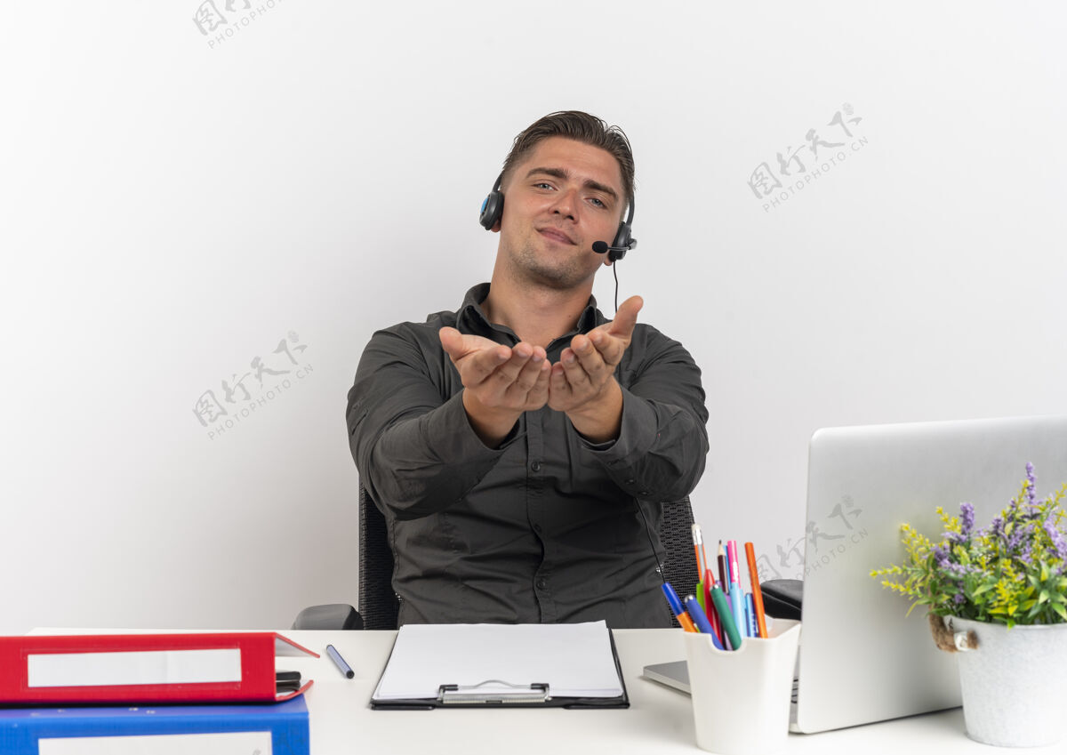 使用年轻而快乐的金发上班族戴着耳机坐在办公桌旁 拿着办公工具 用笔记本电脑 双手分开放在白色背景上 留着复印空间请笔记本电脑坐着
